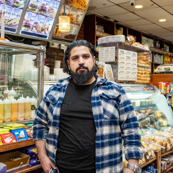 A man with a beard and plaid shirt stands in a deli with food displays and menus.