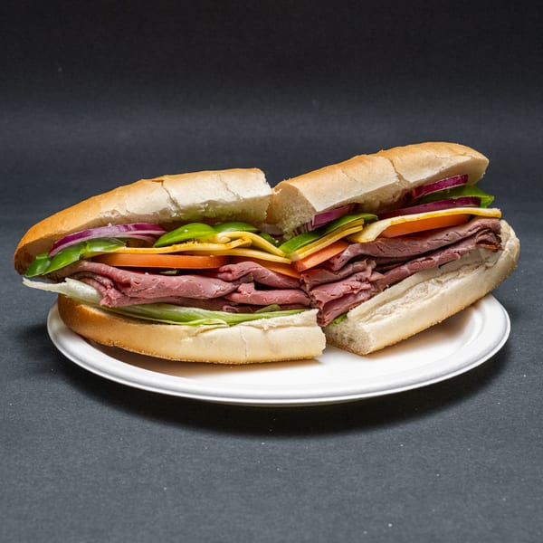 Roast beef sub sandwich, cut in half, with lettuce, tomato, onion, and cheese on a white plate.