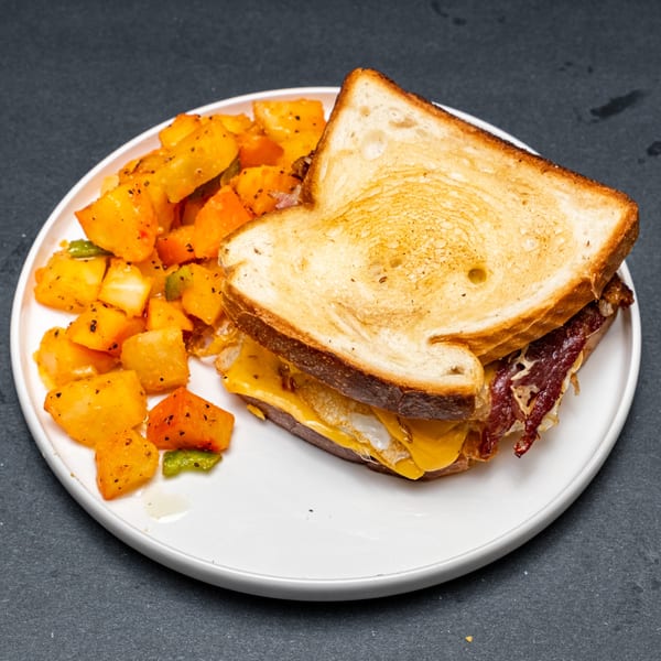 A grilled cheese sandwich with bacon and a fried egg, served with roasted potatoes on a white plate.