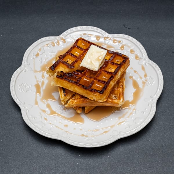 Stack of golden-brown waffles with melting butter and maple syrup on a white decorative plate.
